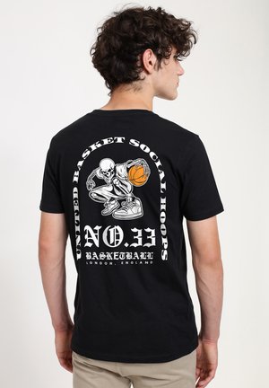 Ung mand der viser en sort T-shirt med en hvid skelet, der dribler en orange basketball, og hvidt stiliseret tekst på ryggen.