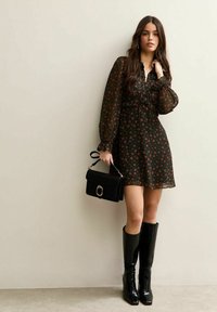 Robe mini noire à fleurs avec des manches bouffantes, taille cintrée et tissu transparent. Associée à des bottes noires montant jusqu'aux genoux et un petit sac à main noir.