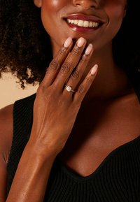 Zilveren ring met een ronde, sprankelende steen, gedragen op een hand met lichtroze nagels. De huidtint is warm en soepel, tegen een gestructureerde zwarte top.