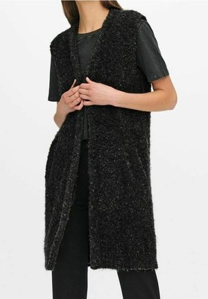 Kobieta nosząca czarną, bezrękawnikową, futrzaną kamizelkę do kolan, na krótkim, czarnym rękawku i czarnych spodniach, trzymając kamizelkę z przodu zapiętą.