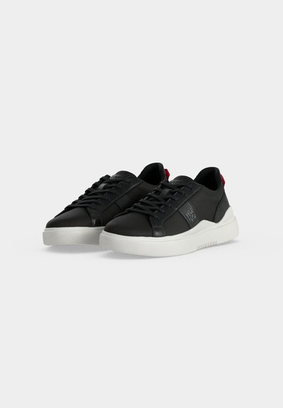 Paire de baskets basses noires avec semelles blanches, lacets noirs et une petite languette rouge au talon, présentée sur fond blanc.