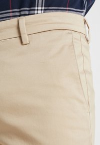 Chino in cotone beige con una texture liscia, design frontale piatto e una tasca laterale con cucitura nascosta.