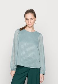 Blusa teal clara com mangas longas e transparentes, apresentando riscas metálicas verticais e um decote redondo. Combinada com calças verdes escuras.