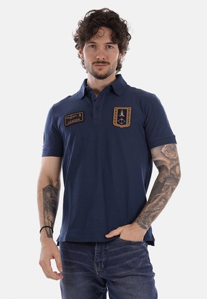 Uomo con capelli ricci che indossa una polo blu navy con toppe etichettate "Leader" e "TROOP TRICOLOR", braccia tatuate, una mano nella tasca dei jeans.