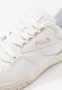 Sneakers bianchi in pelle con lacci in tessuto. Presentano un logo in rilievo prominente, perforazioni per la traspirabilità e una suola testurizzata.