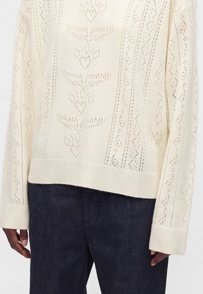 Pull en maille crème avec motifs floraux et géométriques en dentelle porté avec un pantalon foncé, montrant le torse et les bras.