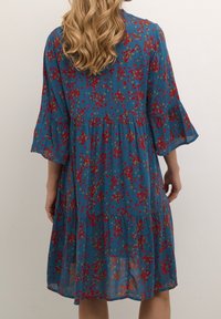 Robe à fleurs en tissu bleu, avec des motifs rouges et verts, des manches évasées, un design à volants et une coupe décontractée, atteignant une longueur mi-mollet.