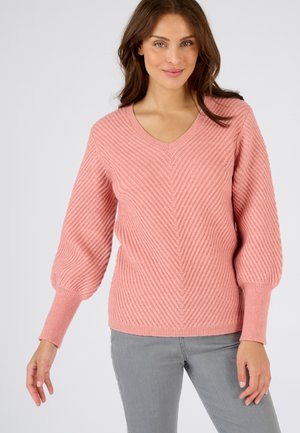 Roze gebreide trui met V-hals, voorzien van diagonale ribpatronen en ballonmouwen. Gepaard met lichtgrijze broeken.