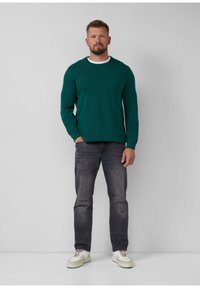 Grüner Pullover mit langen Ärmeln, rundem Halsausschnitt, kombiniert mit dunklen Jeans und weißen Sneakers. Auffällige Details sind leichte Abnutzungen an den Jeans.