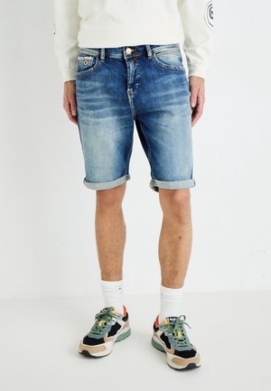 Personne portant un short en denim bleu retroussé, des chaussettes blanches montantes et des baskets multicolores, debout sur un fond blanc uni.