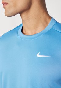 Heledsinine spordipaita, ümarate kraega, valmistatud siledast kangast. Esiküljel on väike valge Nike logo.