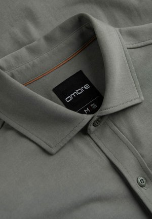 Graues Poloshirt mit einem Kragen, zwei Knöpfen und einer braunen Akzentuierung am Dekolleté. Etikett sichtbar mit Marken- und Größeninformationen. Weiche Stofftextur.