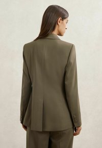 Moteris iš nugaros, dėvinti alyvuogių žalią kostiuminį švarką ir atitinkančias kelnes, stovinti priešais paprastą šviesų foną.