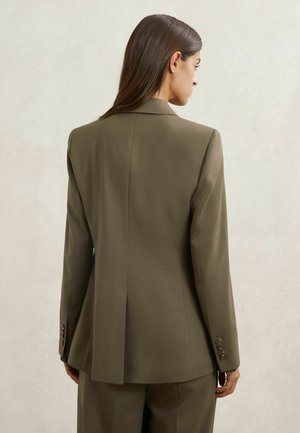 Vrouw van achteren, gekleed in een olijfgroene colbert en bijpassende broek, tegen een effen lichte achtergrond.