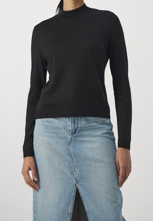 Personne portant un haut noir à manches longues et col montant associé à une jupe en jean bleu clair avec une fente à l'avant.