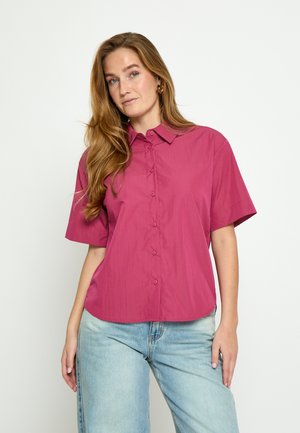 Frau mit langen, blonden Haaren, die ein magentafarbenes kurzärmeliges Hemd mit Knöpfen und hellblaue Jeans vor einem schlichten weißen Hintergrund trägt.