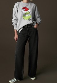 Sweat-shirt gris avec un motif coloré du Grinch portant un chapeau de Père Noël rouge, accompagné d'un jean noir ample et de baskets argentées.