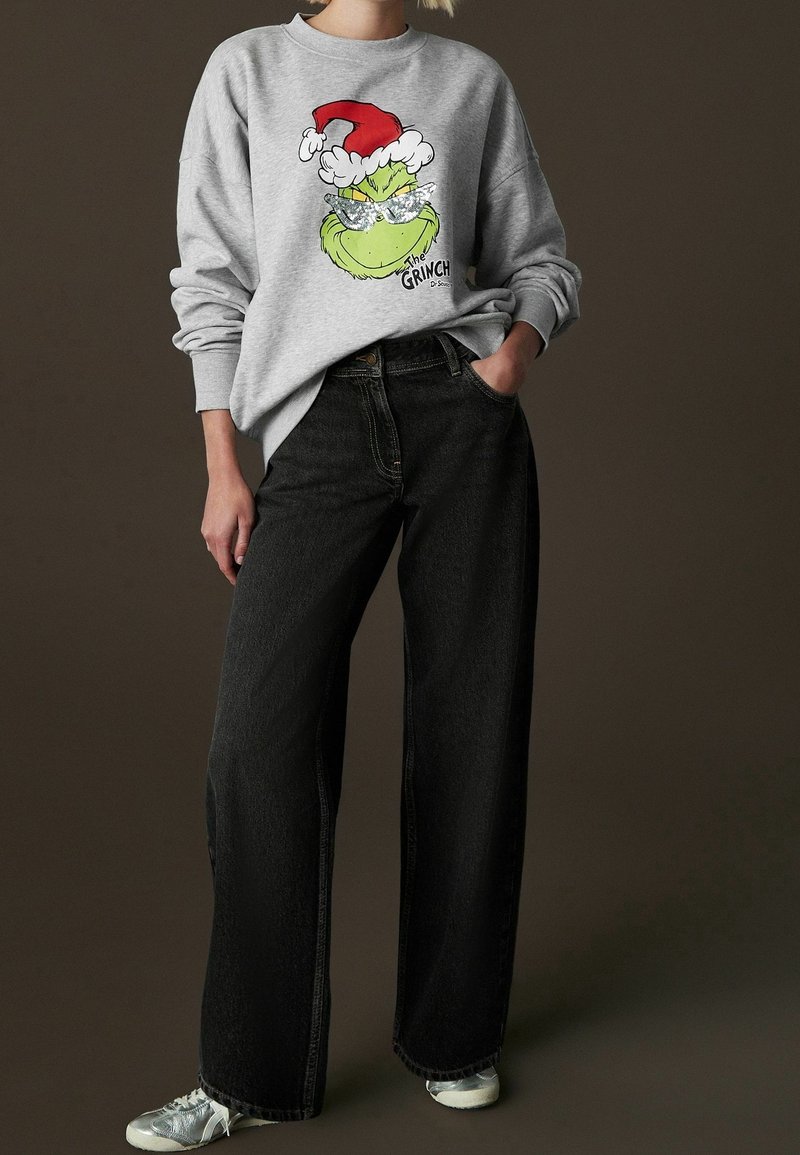 Sweat-shirt gris avec un motif coloré du Grinch portant un chapeau de Père Noël rouge, accompagné d'un jean noir ample et de baskets argentées.