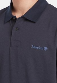 Camisa polo de color azul marino hecha de tela texturizada, con cuello, tapeta de dos botones y logo bordado de Timberland en color azul claro en el pecho.