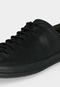 Zapatilla baja de cuero negro con cordones negros y un diseño de puntera lisa y redondeada sobre una suela negra, sobre un fondo sencillo.