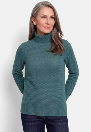 Vrouw met lang grijs haar met een getextureerde teal-coltrui en donkere spijkerbroek, staand tegen een effen lichte achtergrond.