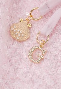 Γουράκι από χρυσό κοχύλι με μαργαριτάρια και χρυσό γράμμα G με στρας, και τα δύο δεμένα σε απαλές ροζ υφασμάτινες κορδέλες σε ροζ δαντελένιο φόντο.