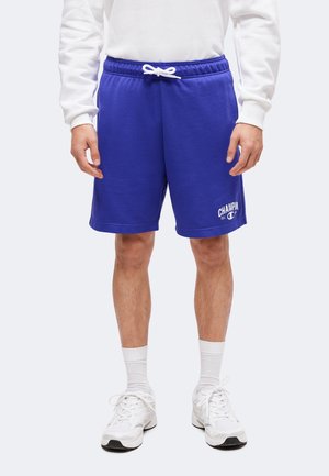 Mann trägt blaue Shorts mit Kordelzug und weißem Champion-Logo, weißes Langarmshirt, weiße Socken und weiße Sneakers und steht vor einem schlichten Hintergrund.
