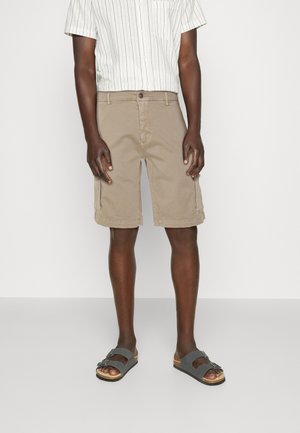 Shorts cargo beiges avec poches latérales, en tissu mélange de coton. Associés à une chemise à manches courtes rayée et des sandales grises.