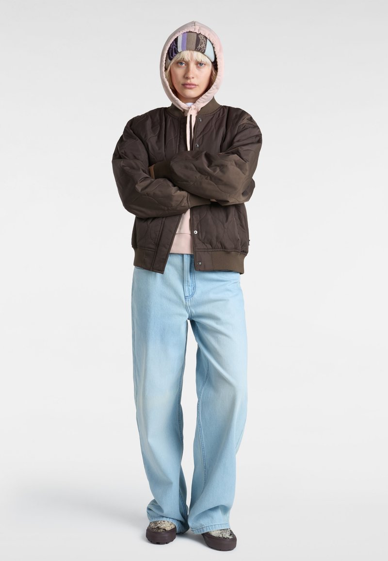 Giacca bomber trapuntata marrone sopra un pantalone con cappuccio rosa chiaro, abbinata a jeans a gamba larga azzurri e sneakers con fantasia.