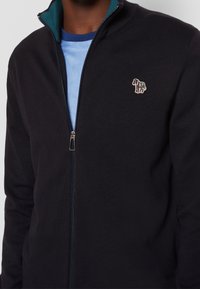 Persona que lleva puesto un suéter negro con cremallera y un pequeño parche de cebra multicolor en el lado izquierdo del pecho, sobre una camisa de color azul claro.