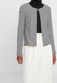 Cardigan a righe blu navy e bianco, realizzato in tessuto strutturato, con scollo rotondo e motivo aperto sul davanti, abbinato a pantaloni bianchi a vita alta.