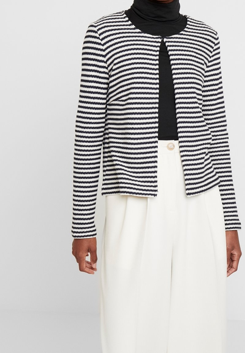 Cardigan a righe blu navy e bianco, realizzato in tessuto strutturato, con scollo rotondo e motivo aperto sul davanti, abbinato a pantaloni bianchi a vita alta.