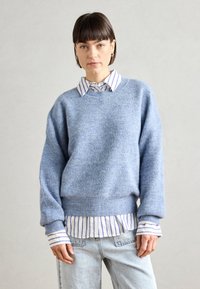 Light blue knit sweater with a ribbed texture, round neckline, and dropped shoulders layered over a white and blue striped shirt and jeans.