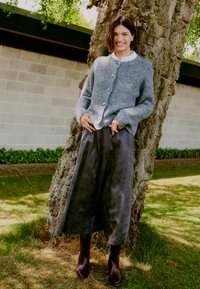 Cardigan en tricot gris avec de grands boutons, associé à une jupe sombre et fluide et des bottes en cuir marron jusqu'au genou, sur fond d'arbre.