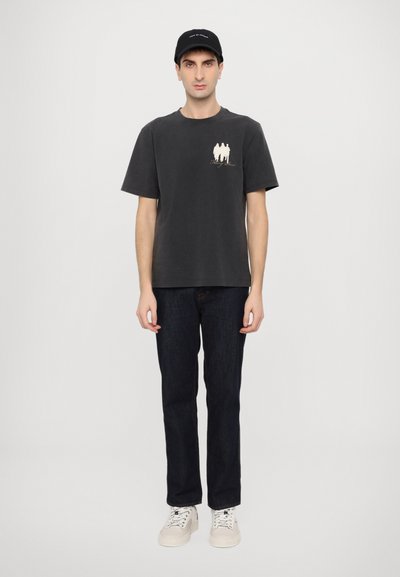 Black cotton t-shirt with short sleeves, featuring a graphic on the chest, paired with dark denim jeans and white sneakers. Black cap on head.
