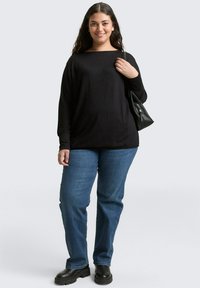 Zwarte lange mouw top met een brede halslijn, gecombineerd met blauwe rechte spijkerbroek en zwarte platformlaarzen. Model houdt een zwarte handtas vast.