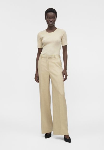 Γυναίκα με μπεζ ριμπ πλεκτό κοντομάνικο τοπ και μπεζ παντελόνια με φαρδιά μπατζάκια ψηλόμεσα, καθώς και μαύρα μυτερά παπούτσια, στέκεται μπροστά σε λευκό φόντο.