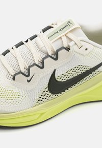 Chaussure de course légère avec une tige en maille respirante de couleur crème et verte, présentant des accents gris foncé et un emblème noir caractéristique.