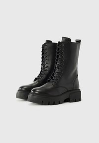 Botas de tobillo de cuero negro con suela de goma gruesa, puntera redonda, ojales de metal y cierre de cordones en la parte delantera. Incluye una lengüeta en la parte posterior.