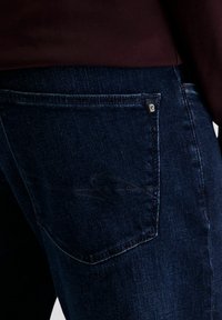 Donkerblauwe spijkerbroek met een achterzak voorzien van een klein logolabel. De stof heeft een gladde textuur met subtiele vervagingen.