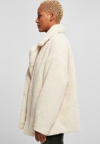 Overmaat crèmekleurige nepbontjas met een brede kraag, zachte textuur en een relaxed fit, met grote mouwen en zonder zichtbare sluitingen.