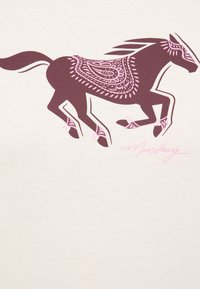 Dunkelbraunes Pferde-Graphic mit rosa dekorativen Mustern, auf einem hellen cremefarbenen Shirt gedruckt. Unten befindet sich eine Unterschrift in Rosa.
