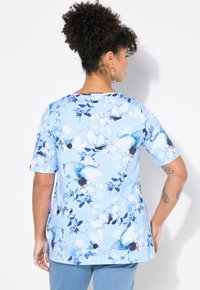 Blusa floral en azul claro con mangas cortas, fabricada en un tejido suave y con un diseño de flores abstractas en azul oscuro y blanco. Cuello redondeado con aberturas laterales.