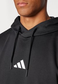 Črna kapuca z mehkobo teksture, na kateri je na sprednji strani bel logotip Adidas. Vrvica za zategovanje je vidna pri ovratniku.