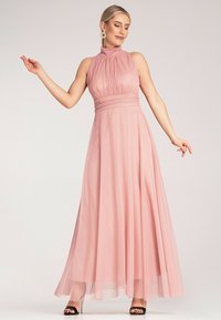 Różowa maxi sukienka na szyję wykonana z lekkiego materiału, z plisami, dopasowanym talią i zwiewnym dołem. Noszona z czarnymi sandałami na obcasie.