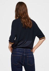 Haut en maille bleu marine avec un design en blocs de couleurs, présentant un col côtelé à rayures orange, associé à un jean denim foncé, vu de dos.