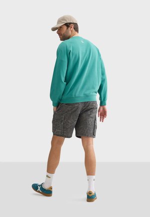 Turquoise sweatshirt, grijze gemusterde shorts, blauwe sneakers, beige pet. Casual sportieve stijl met zichtbare stiksels en zakken.