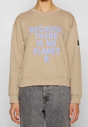 Beżowa bluza z jasnoniebieskim napisem "Because there is no planet B", noszona z szarymi dżinsami, pokazana od szyi do bioder.