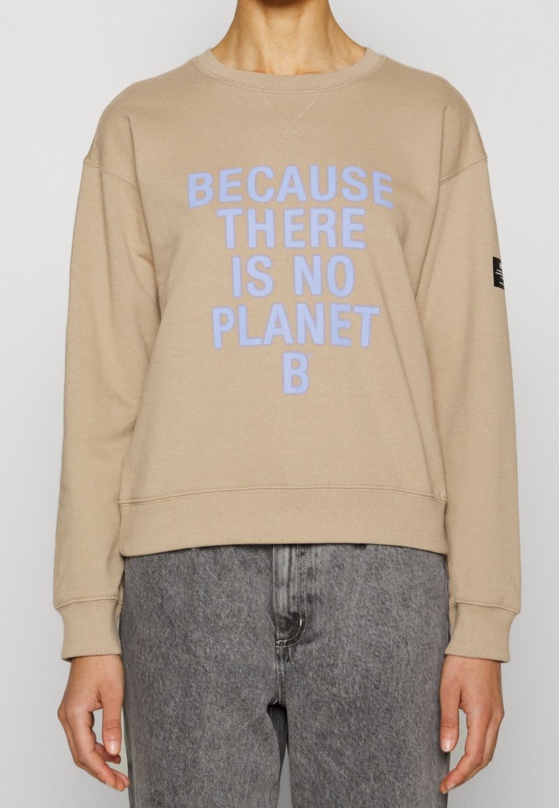 Sweat beige avec texte bleu clair "Because there is no planet B", porté avec un jean gris, visible du cou jusqu'aux hanches.