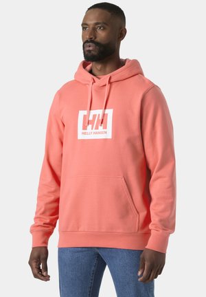 Hombre con sudadera coral Helly Hansen con bolsillo frontal y vaqueros azules, de pie contra un fondo claro y sencillo.